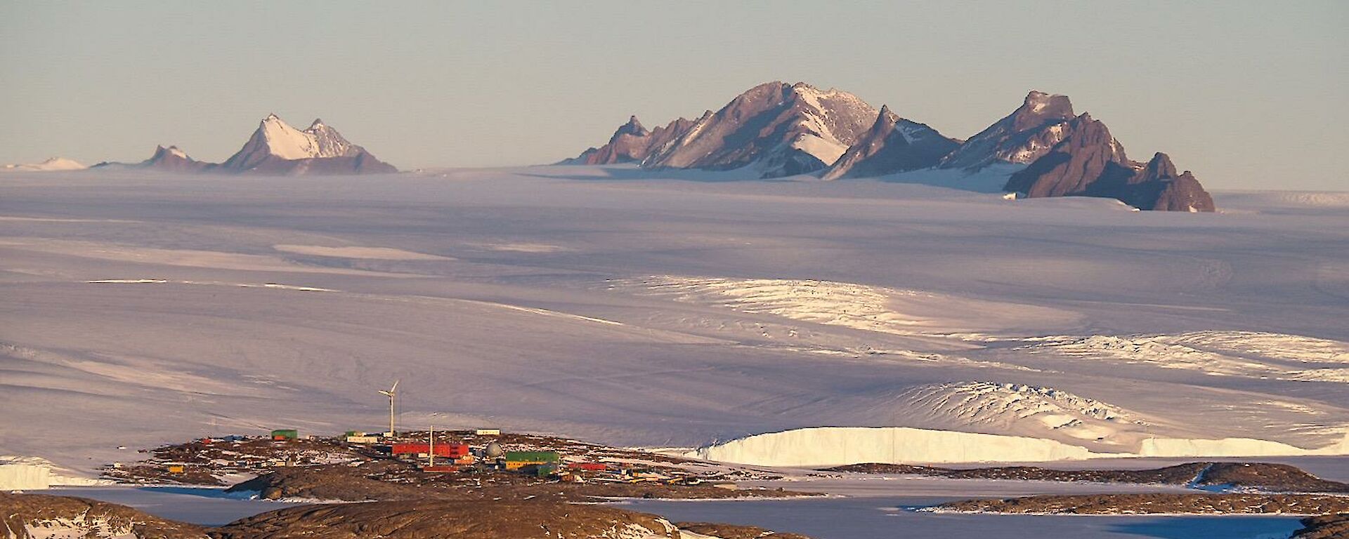 Mawson station seen from a distance with ice plateau behind and snow covered Framnes Mountains in distance