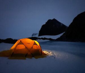 Tent pitched on frozen lake with mountains overlooking. Lights on inside tent make it glow bright orange.