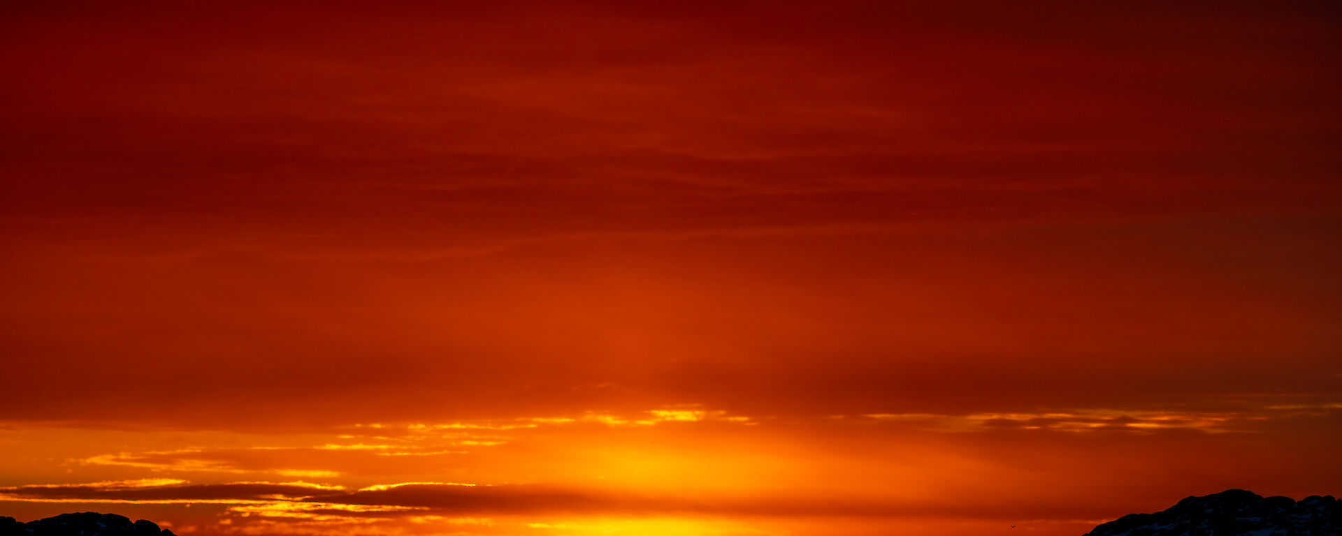 A richly-coloured sunset scene, showing an ocean and distant icebergs in silhouette beneath a darkening vermilion sky. Low clouds above the sunset are outlined in bright gold