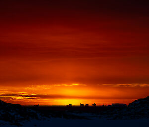 A richly-coloured sunset scene, showing an ocean and distant icebergs in silhouette beneath a darkening vermilion sky. Low clouds above the sunset are outlined in bright gold