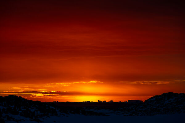 A richly-coloured sunset scene, showing an ocean and distant icebergs in silhouette beneath a darkening vermilion sky. Low clouds above the sunset are outlined in bright gold