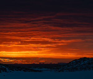 A richly-coloured sunset scene. Stacked layers of low clouds are lit from beneath in fiery colours above a dark ocean