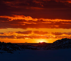 The sun setting behind an iceberg on the ocean. Low clouds massed above a clear horizon contrast dark red with light amber. The snow-covered ground in the foreground is in purplish shadow