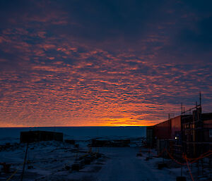 A sunrise scene with sheds, scaffolding, poles and ropes in the shadowed foreground. Above is a "mackerel sky" of fine-grained cloud, dark violet stippled with bright coral highlights over an orange glow where the sun is about to rise