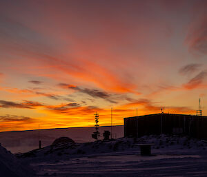 A sunrise scene over a large shed. Streaks of high cloud above the sunrise are lit with flame-like colours, while lower wisps of cloud appear dark and smoky. The snowy foreground, in shadow, has a violet tint