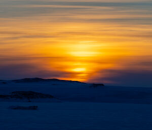 A sunrise scene over an iced ocean and snowy ground. The sky is full of thick, high clouds that seem to stretch and expand the sun's glow in a wavering manner, like a bright reflection on dark water. The snow and ice on the ground, in silhouette, appear dark blue in contrast with the golden sunlight