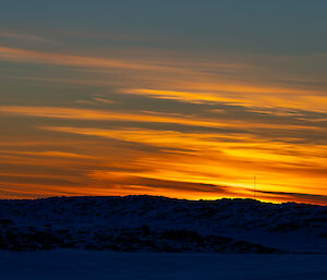 A sunrise scene over rocky, snowy ground with three radio masts rising in the distance. The dawn sky appears deep greenish-grey, invaded by fiery gold and red stripes of high cloud. The ground appears dark blue in contrast