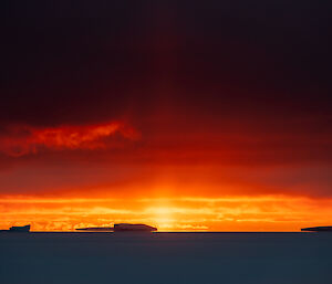 Icebergs in an ocean sheeted over with ice, in rich sunset contrasts of light, shade and colours. The ice appears dark grey-blue, with shreds of low cloud lit bright yellow in the clear orange sky at the horizon. Higher up where thick cloud dominates the sky, the colours fade through scarlet into darkness