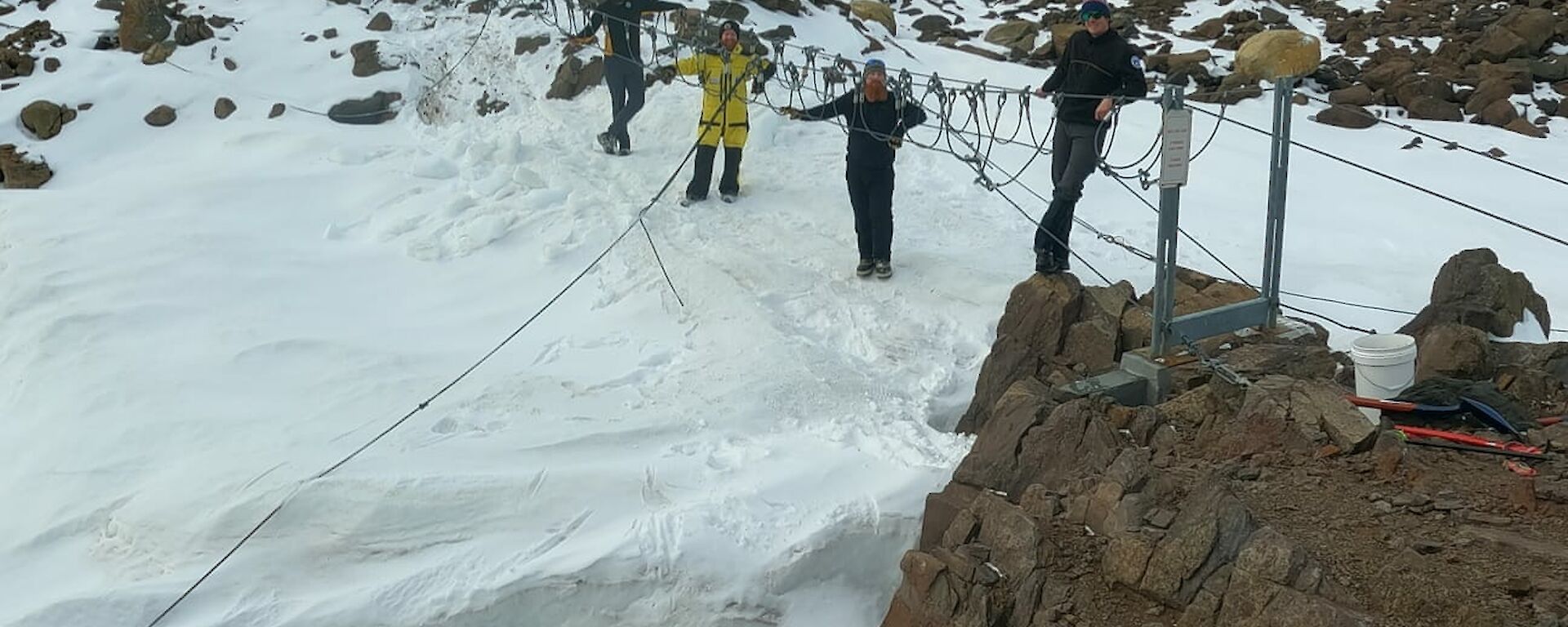 A group of men standing around a suspension bridge over snow and rocks