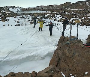 A group of men standing around a suspension bridge over snow and rocks