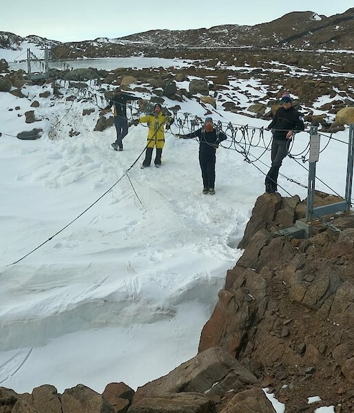 A group of men standing around a suspension bridge over snow and rocks