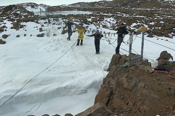 A group of men standing around a suspension bridge over snow and rocks
