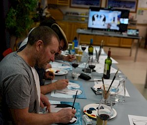 Man in foreground sitting at table and painting, in the distance a TV screen with instructional video