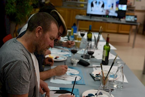 Man in foreground sitting at table and painting, in the distance a TV screen with instructional video