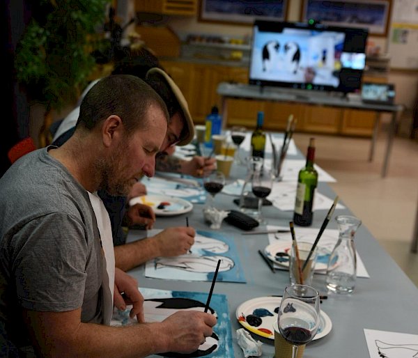 Man in foreground sitting at table and painting, in the distance a TV screen with instructional video