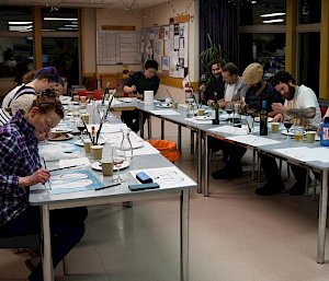 Tables set in U shape, people sitting around outside painting on sheets on table before them, with paints and brushes laid out