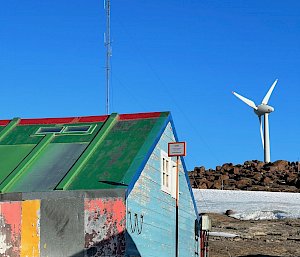 In foreground a timber clad hut painted in mulit-colours and in the background a large wind turbine