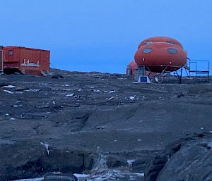 Rocky ground in foreground, blue sky above. On the horizen are huts, one orange square and one orange melon shaped