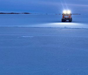 Twilight colours of blue-purple of sea-ice that merges into horizon and sky. in middle distance an orange hagglunds vehicle with roof mounted headlights on.