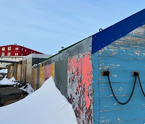 In foreground the side of timber clad hut painted multi colours, in the distance elevated is large red building