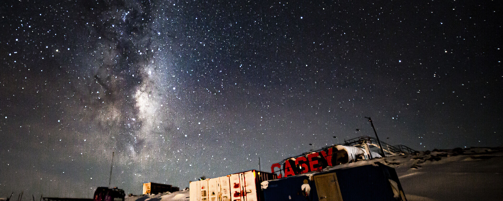 A night scene in a location with shipping containers. Large, red letters spelling "CASEY" are erected on a rise behind them. The night sky is thickly- and brightly-starred and the pale glow of the Milky Way can be seen extending from the horizon