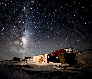 A night scene in a location with shipping containers. Large, red letters spelling "CASEY" are erected on a rise behind them. The night sky is thickly- and brightly-starred and the pale glow of the Milky Way can be seen extending from the horizon