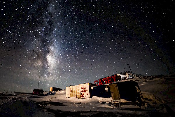 A night scene in a location with shipping containers. Large, red letters spelling "CASEY" are erected on a rise behind them. The night sky is thickly- and brightly-starred and the pale glow of the Milky Way can be seen extending from the horizon