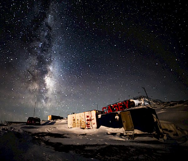 A night scene in a location with shipping containers. Large, red letters spelling "CASEY" are erected on a rise behind them. The night sky is thickly- and brightly-starred and the pale glow of the Milky Way can be seen extending from the horizon