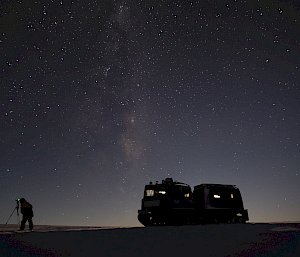 A night scene with a Hägglunds vehicle parked on a plain. A man is standing to the left, looking through a camera set up on a tripod. The sky is densely scattered with stars and the faint glow of the Milky Way is visible
