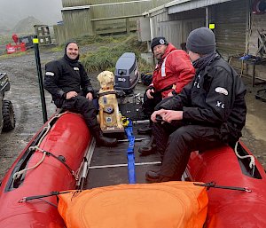 Three people and a plastic dog sit in a red rubber boat onshore