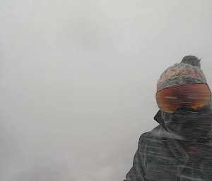 Torso and head of person, completely covered with neck warmer, goggles and beanie, with snow whipping around them and nothing to view in the background