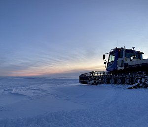 A snow groomer vehicle smoothing out an area on a wide, snowy plain under a twilight sky