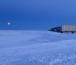 A number of large transport containers mounted on skis, parked in a row on a snowy plain. A golden full moon is rising in the background