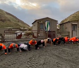 A group of people doing push ups on the grey sand in front of two wooden buildings
