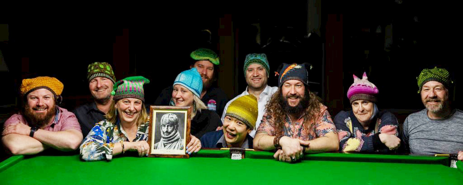 A group of people leaning against a pool table wearing beanies and smiling. One woman is holding a framed picture of explorer Douglas Mawson