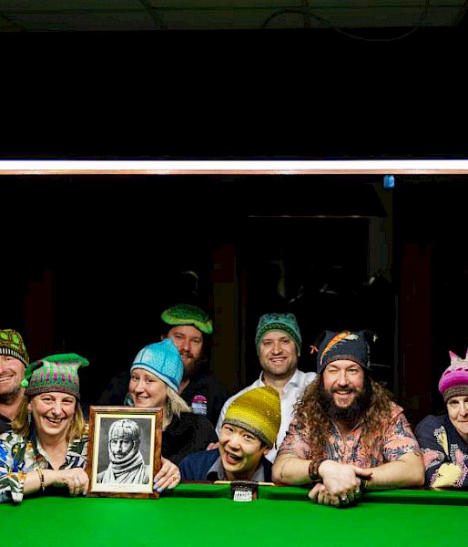 A group of people leaning against a pool table wearing beanies and smiling. One woman is holding a framed picture of explorer Douglas Mawson