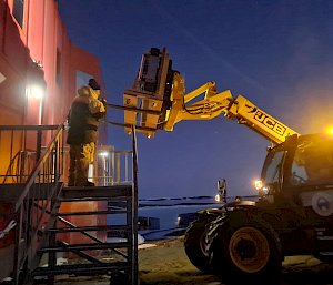 JCB forklift lifts base of treadmill in wooden pallet onto balcony which is entry to the Red Shed (Living Quarters)