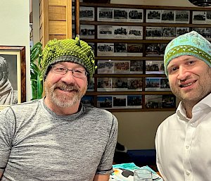 Two men wearing blue/green beanies are smiling at the camera, behind them is a picture of Sir Douglas Mawson wearing his own knitted balaclava