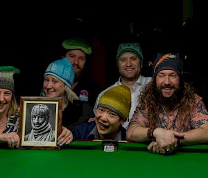10 expeditioners, leaning over pool table, wearing the 10 hats from Hadley's project and holding the portrait of Sir Douglas Mawson wearing his knitted beanie