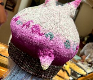 Close up photo of a beanie worn by a woman, from behind. Beanie is shaded from dark pink at bottom to light pick at top, and has two knitted horns rising from the top and one at bottom back pointing downwards