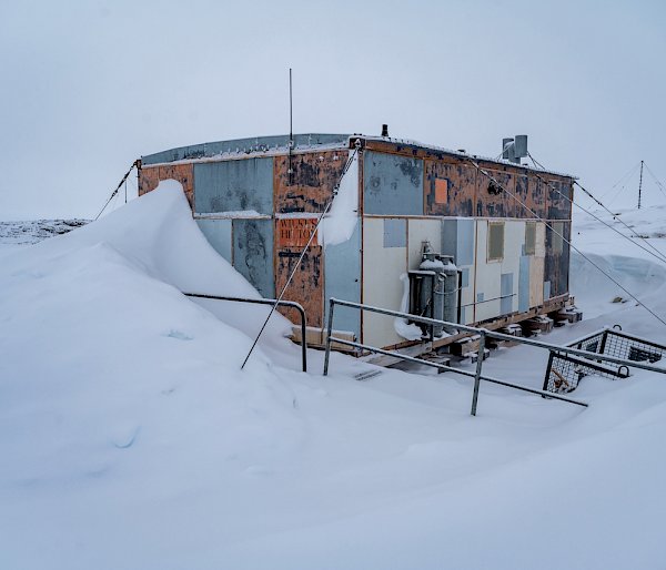 A rectangular hut in a snowy location. The hut's exterior is made of weathered wood and metal panels. A tall pile of snow has built up against its door, blocking entry