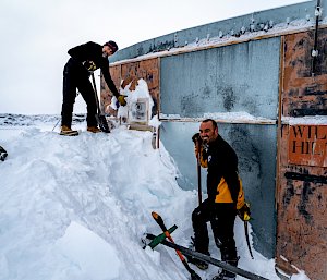 Two men standing on the massive snow pile, ready to start digging it away with shovels. One man gestures towards the sign visible above the snow-blocked door, which reads "Hilton Wilkes"