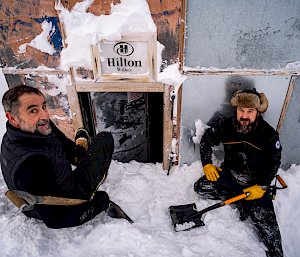 Two men with shovels sitting on the massive snow pile in front of the hut door. Enough snow has been dug away to reveal the top half of the door