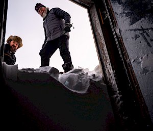 From inside the hut, the door opens onto an apparent wall of snow half as tall as the doorway. Two men stand on top of this 'snow wall', looking down into the hut