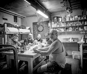 A black and white photo of five men seated at the kitchen table with playing cards and wine glasses, smiling at the camera