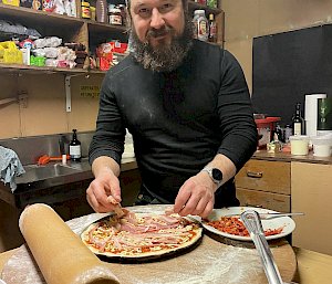 A man is laying toppings on a pizza on top of a wooden board on the kitchen table. A large rolling pin, a container of pizza sauce and a plate of sun-dried tomatoes sit nearby