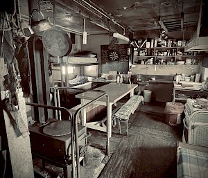 An artistically faded photo of the inside of Wilkes hut, showing a worn wooden floor, metal cooking utensils hanging from the ceiling, bunk beds in the corner, a wood stove, a kitchen table with benches and well-stocked kitchen shelves