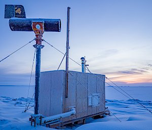 A small wooden hut standing on snowy ground, anchored in place with guywires. A pipe runs out from the hut and connects to a mast, on top of which a horizontally-aligned metal cylinder is fixed, presumably once used for measuring atmospheric conditions