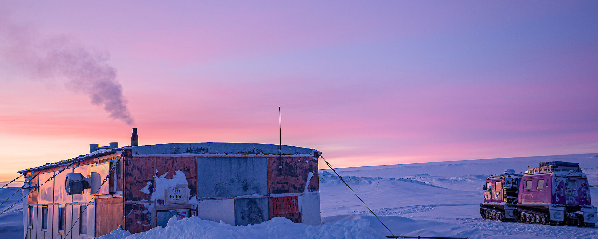 The Wilkes hut in the snow. Sunrise casts pinkish light on high clouds in the sky, contrasting with blue-tinted shadow on the snow. Smoke rises from the hut's chimney. A purple Hägglunds vehicle is parked a short distance from the hut