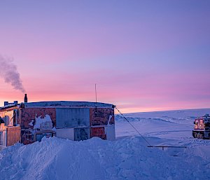 The Wilkes hut in the snow. Sunrise casts pinkish light on high clouds in the sky, contrasting with blue-tinted shadow on the snow. Smoke rises from the hut's chimney. A purple Hägglunds vehicle is parked a short distance from the hut